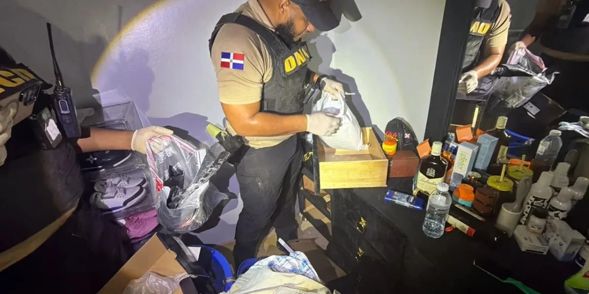 Un agente de la DNCD, con chaleco antibalas y la bandera dominicana en el hombro, revisa una bolsa con evidencias dentro de una vivienda durante uno de los allanamientos en Azua
